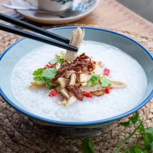 Traditional bubur ayam rice porridge with shredded chicken, fried shallots, and fresh cilantro in white bowl.