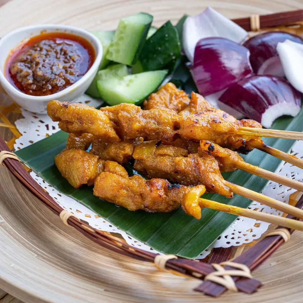 Satay ayam with grilled chicken skewers, peanut sauce, cucumber, and red onions on banana leaf