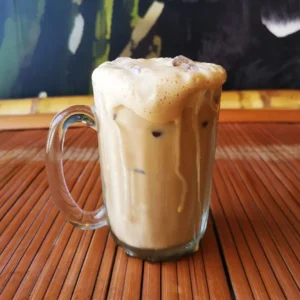 Teh tarik Malaysian pulled tea with creamy milk foam in clear glass mug on wooden bamboo table.
